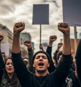 A close-up, ground-level view of a diverse group of determined protestors marching in an urban environment, their faces resolute. Many hands are raised, some clenched into fists, others holding tall, blank protest signs pointing towards a dramatic, cloudy sky.